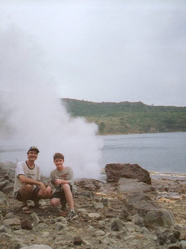 Two people kneel on a rocky terrain near a steaming geothermal vent by a lake, with hills in the background.