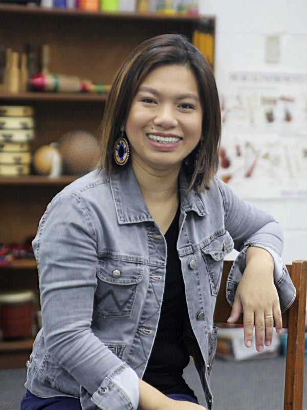 A woman sits indoors wearing a denim jacket and colorful earrings, smiling towards the camera. Shelves with various objects are visible in the background.