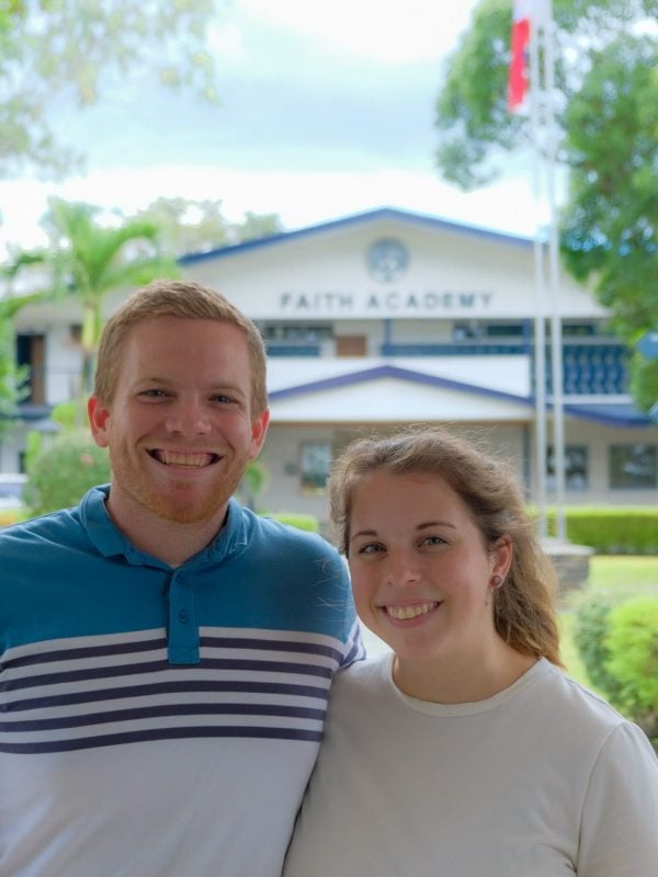 A man and a woman stand side by side, smiling in front of a building labeled "Faith Academy" with a flagpole and trees in the background.