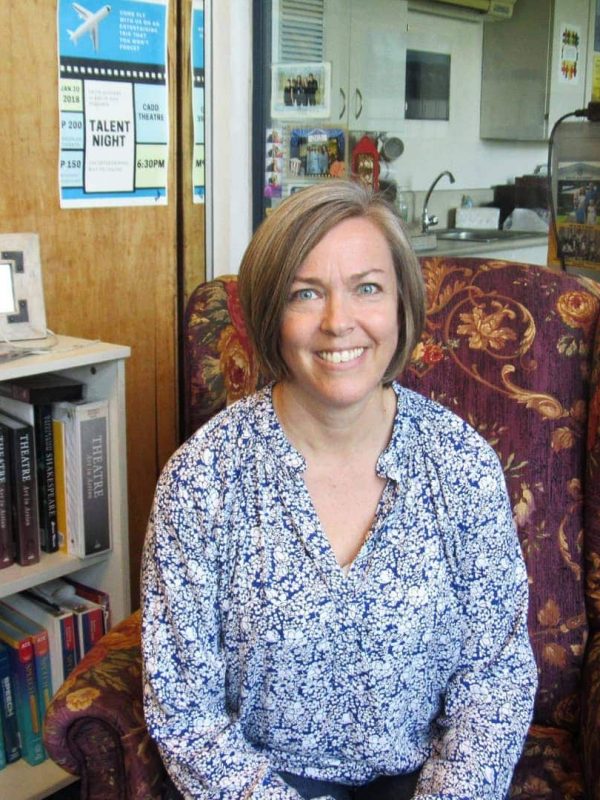 A woman with short hair wearing a blue patterned blouse is sitting on an armchair in a room with bookshelves and posters in the background.