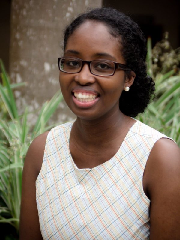 A woman wearing glasses and a white plaid dress is smiling in an outdoor setting with plants and a tree in the background.