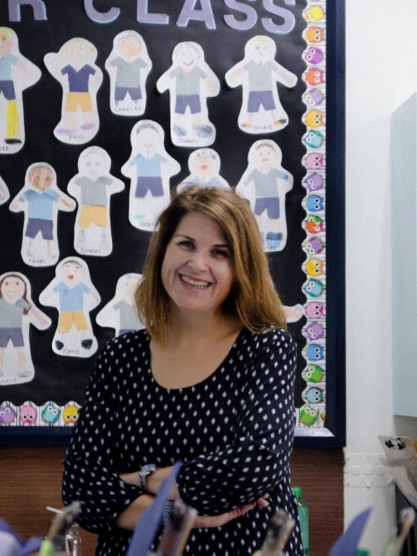 A woman with long brown hair stands smiling in front of a bulletin board displaying children's drawings. The display has a title "Our Class" and two owl decorations labeled January and July.