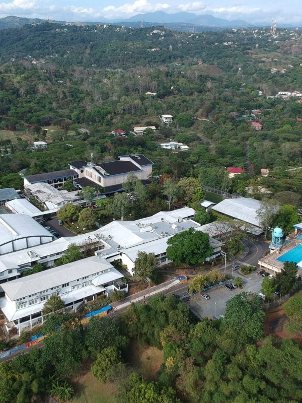 Aerial view of a large campus with multiple white buildings, a swimming pool, sports fields, and surrounding greenery and hills.